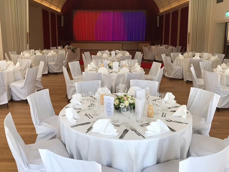 Festlich gedeckte Tische in einem Saal mit Bühne.