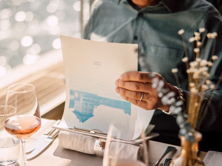 Person in Restaurant hält Menü in der Hand, mit Wasser im Hintergrund.