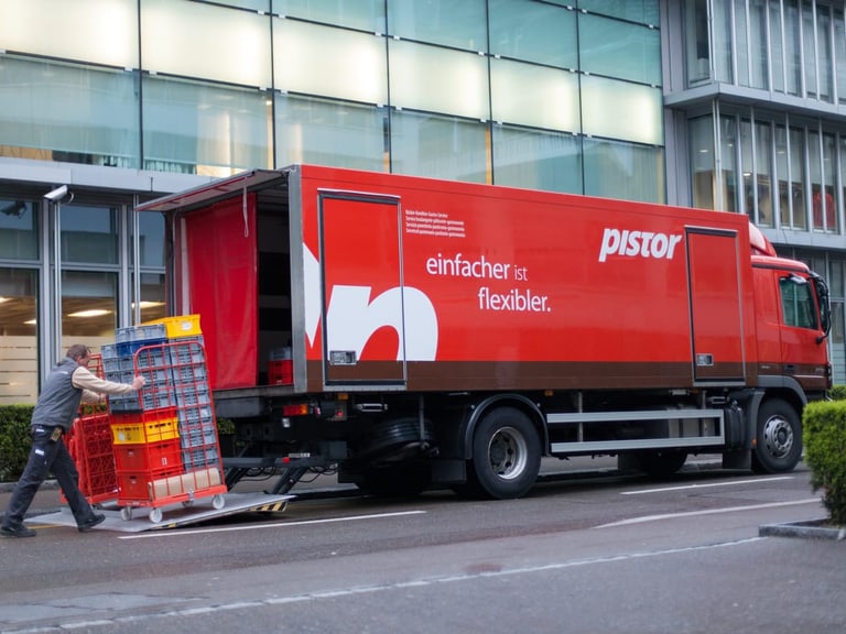Mann lädt Rollcontainer in roten Lastwagen vor Bürogebäude.