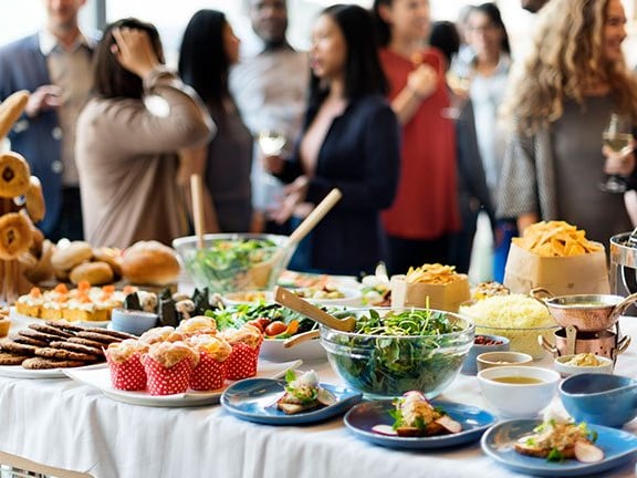 Buffettisch mit verschiedenen Speisen, Menschen im Hintergrund stehend.