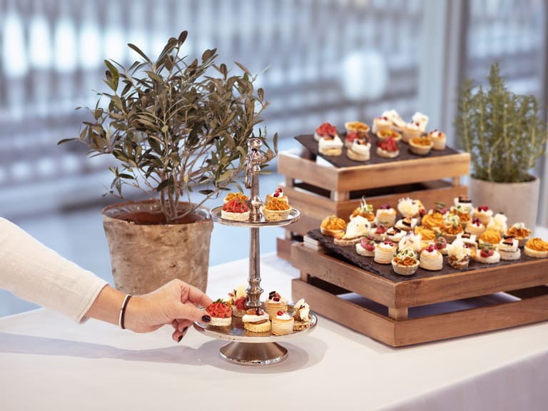 Hand nimmt Häppchen von Tisch mit Pflanze und Tabletts voller Speisen.