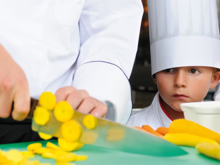 Kind im Kochhut beobachtet Gemüsezubereitung mit gelber Zucchini.