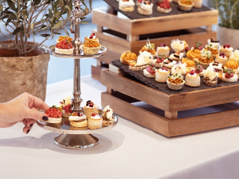 Hand nimmt Häppchen von einer Etagere neben einem Tisch mit Vorspeisen.