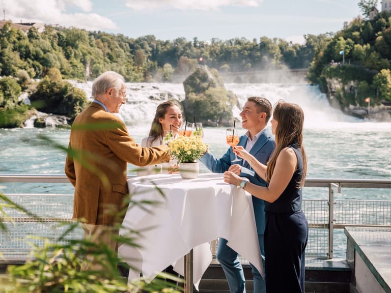 Gruppe von Menschen stossen an einem Stehtisch am Wasserfall an.