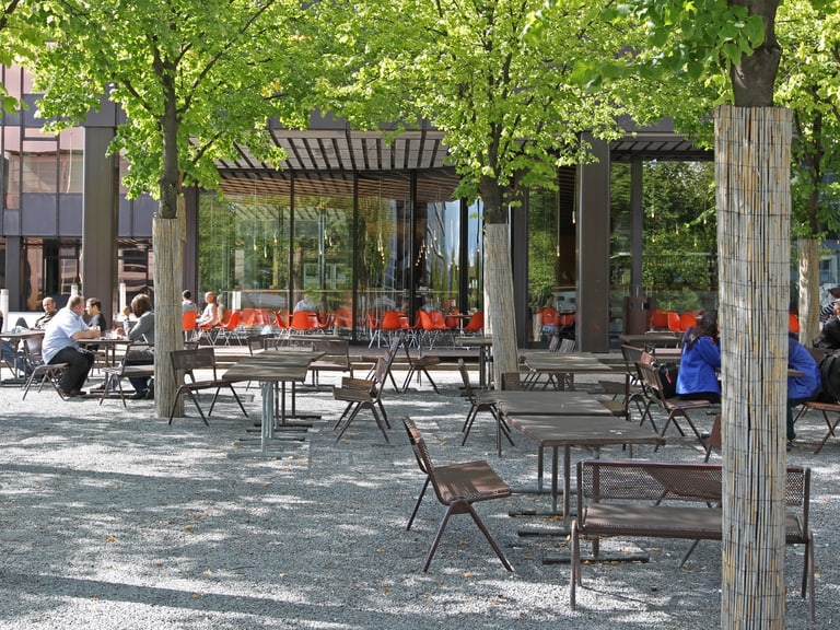 Aussenterrasse mit Tischen und Bäumen, Menschen sitzen im Freien.
