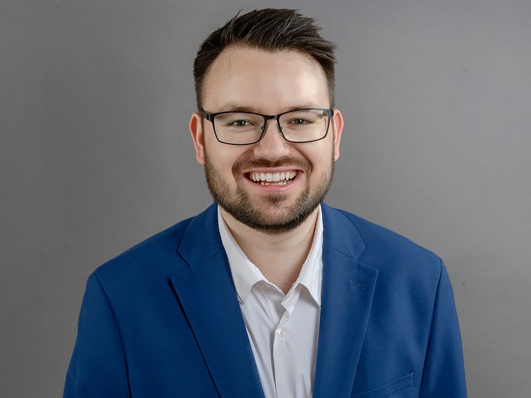 Mann mit Brille in blauem Anzug lächelt vor grauem Hintergrund.