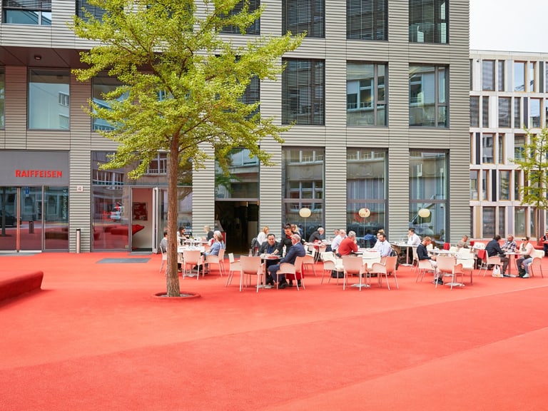 Menschen sitzen im Freien auf roten Flächen vor modernen Gebäuden.