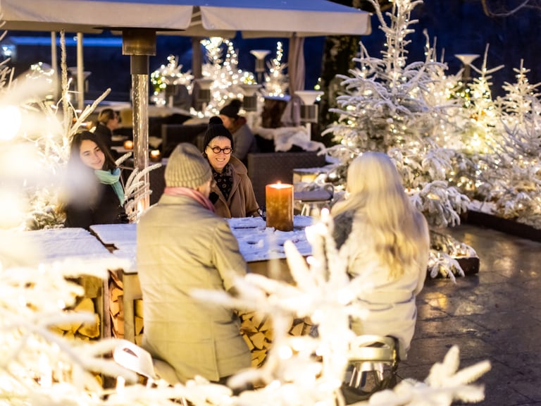 Menschen sitzen an einem Tisch im verschneiten Aussenbereich eines Restaurants mit Lichtern.