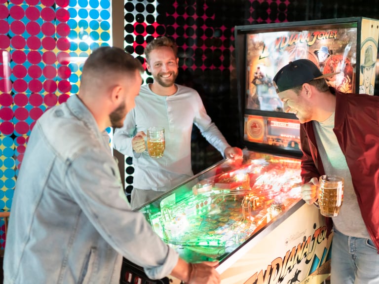 Drei Männer spielen Flipper und halten Bierkrüge.