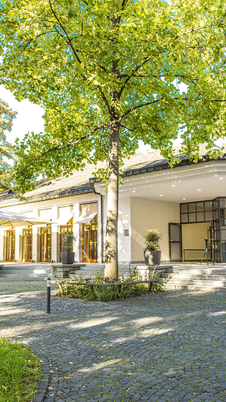 Gebäude mit Bäumen und gepflastertem Vorplatz bei Sonnenschein.