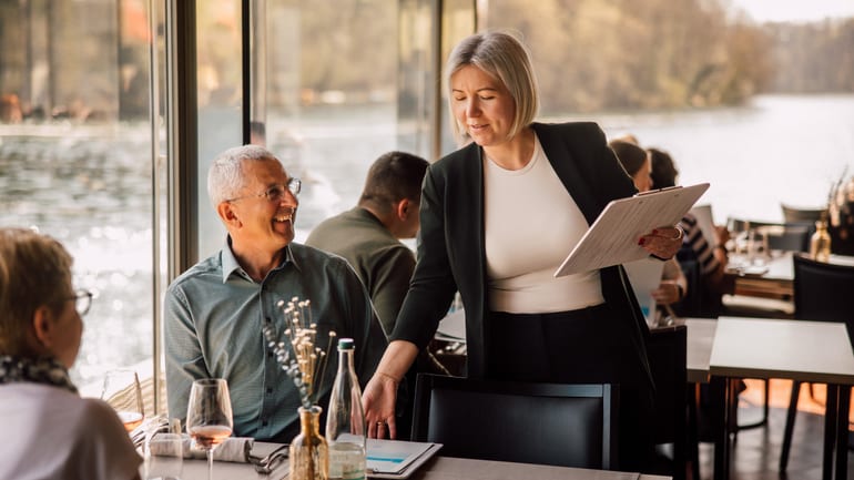 Kellnerin interagiert mit Gästen am Tisch in einem Restaurant am Wasser.