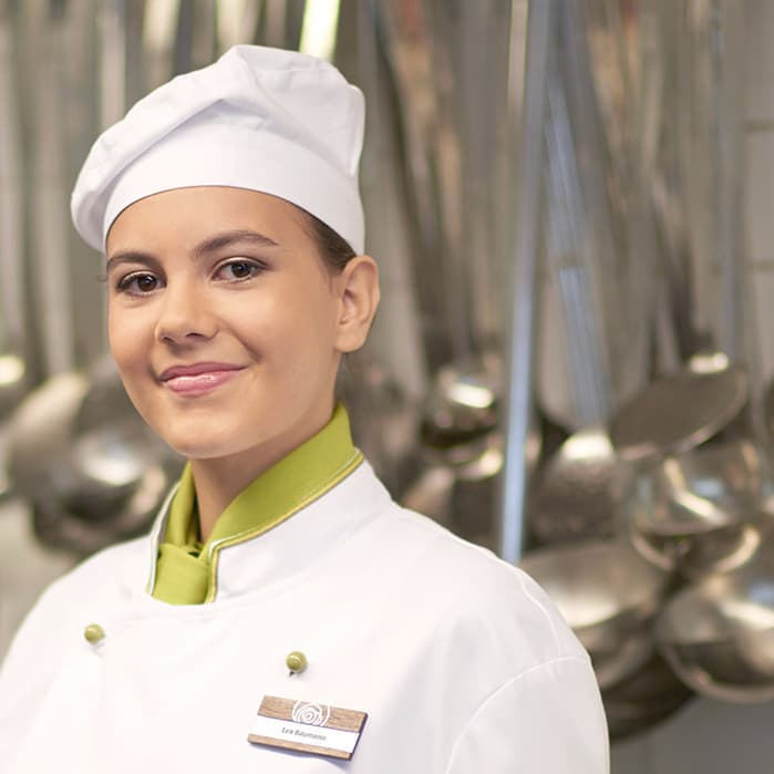 Köchin in weisser Uniform in einer Küche mit Kochutensilien im Hintergrund.